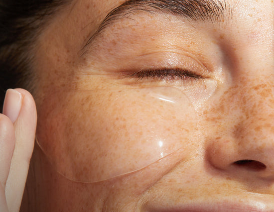 A close-up of a person's face with freckles, focusing on the area around the eye. The person is wearing a Tropci's Hydrogel Eye mask, which covers the under-eye region. The person's eye is closed, and they are gently touching the eye mask with their fingers.